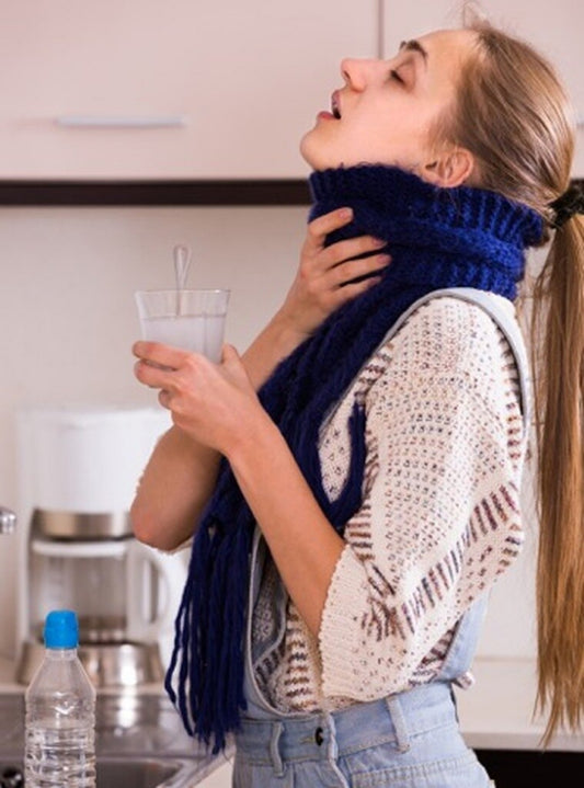 Woman in a kitchen wearing a blue knitted scarf, holding a glass of water and gargling.